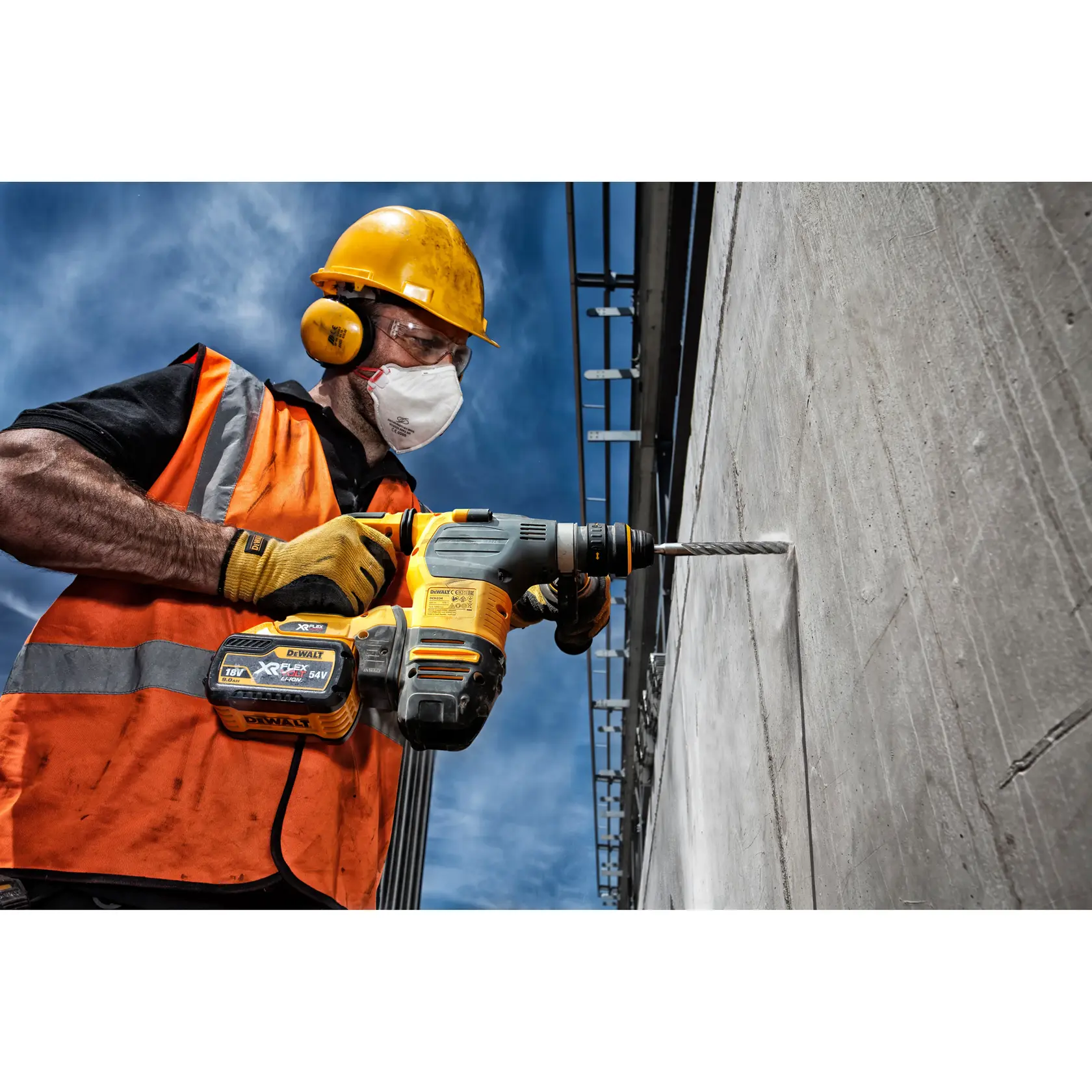 A construction worker wearing a yellow hard hat, ear protection, safety vest, and gloves is using a DEWALT power drill to work on a concrete wall at a construction site under a blue sky.