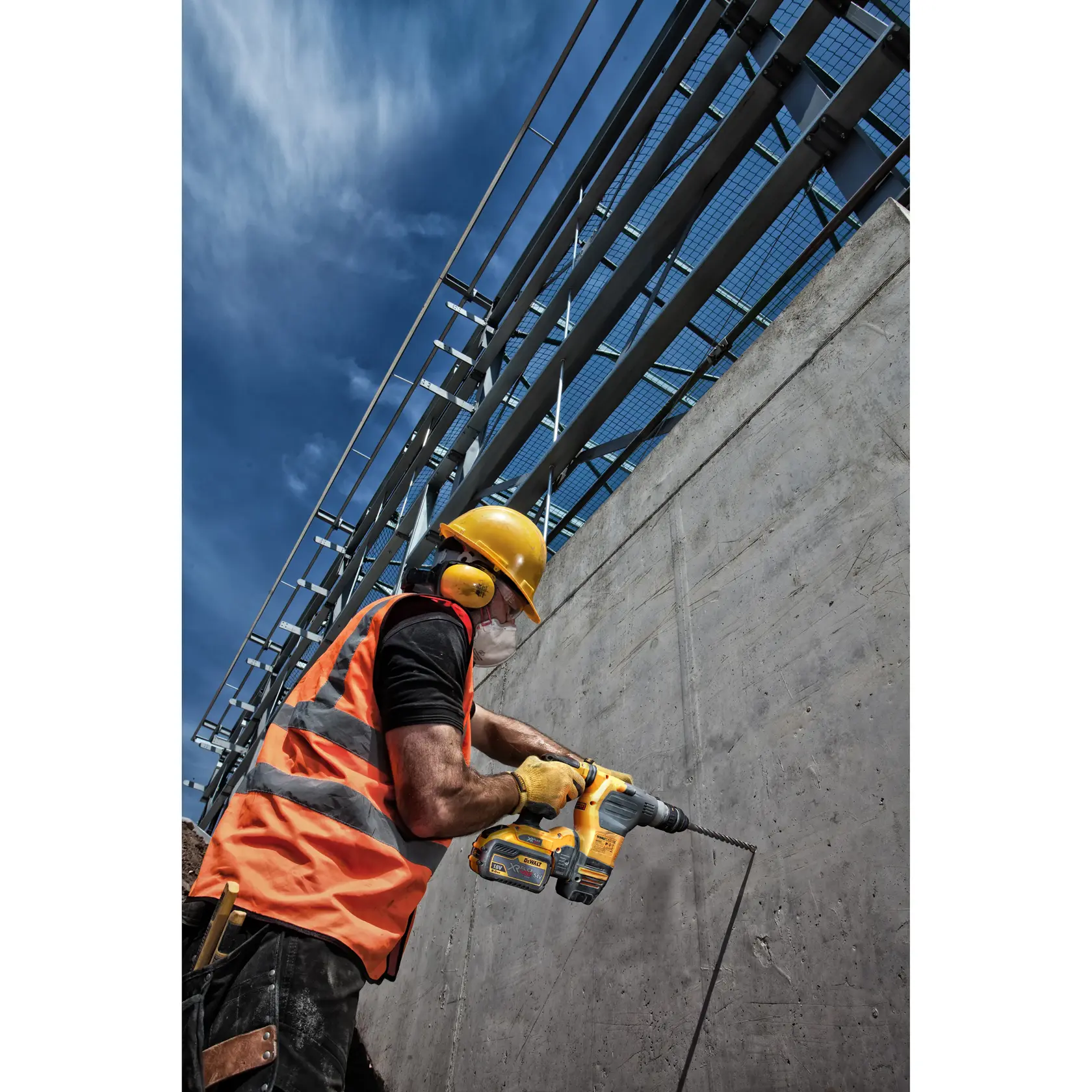 A construction worker wearing a yellow hard hat, orange reflective vest, ear protection, and gloves is using a yellow DEWALT power tool to drill into a concrete wall at an outdoor construction site.