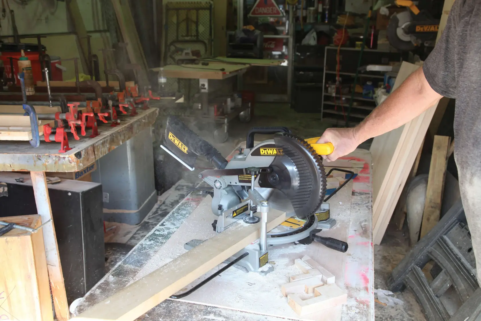 A person operating a DEWALT miter saw (SKU: DW714) to cut a wooden board in a workshop. Wood dust and clamps are visible on the worktable, with various tools and materials in the background.