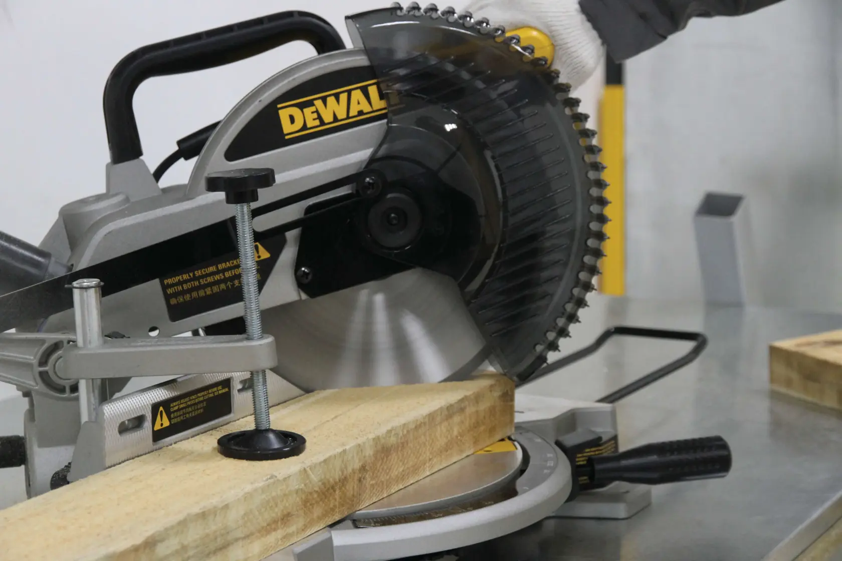 A DEWALT miter saw is being used to cut a piece of wood on a metal workbench. A person wearing a glove is operating the saw.