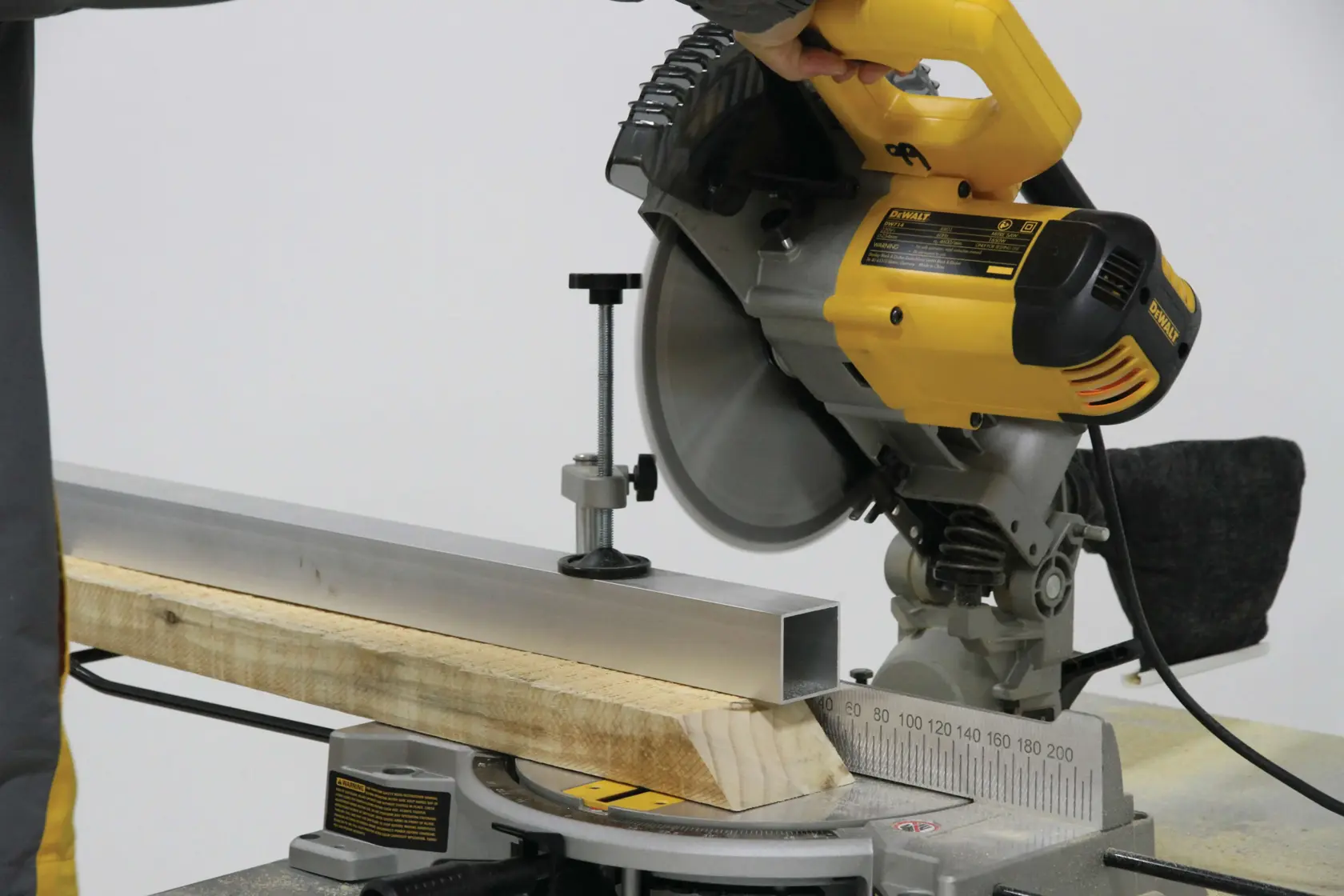 A yellow DEWALT miter saw being used to cut a piece of metal and a piece of wood, held securely in place on the saw’s base.