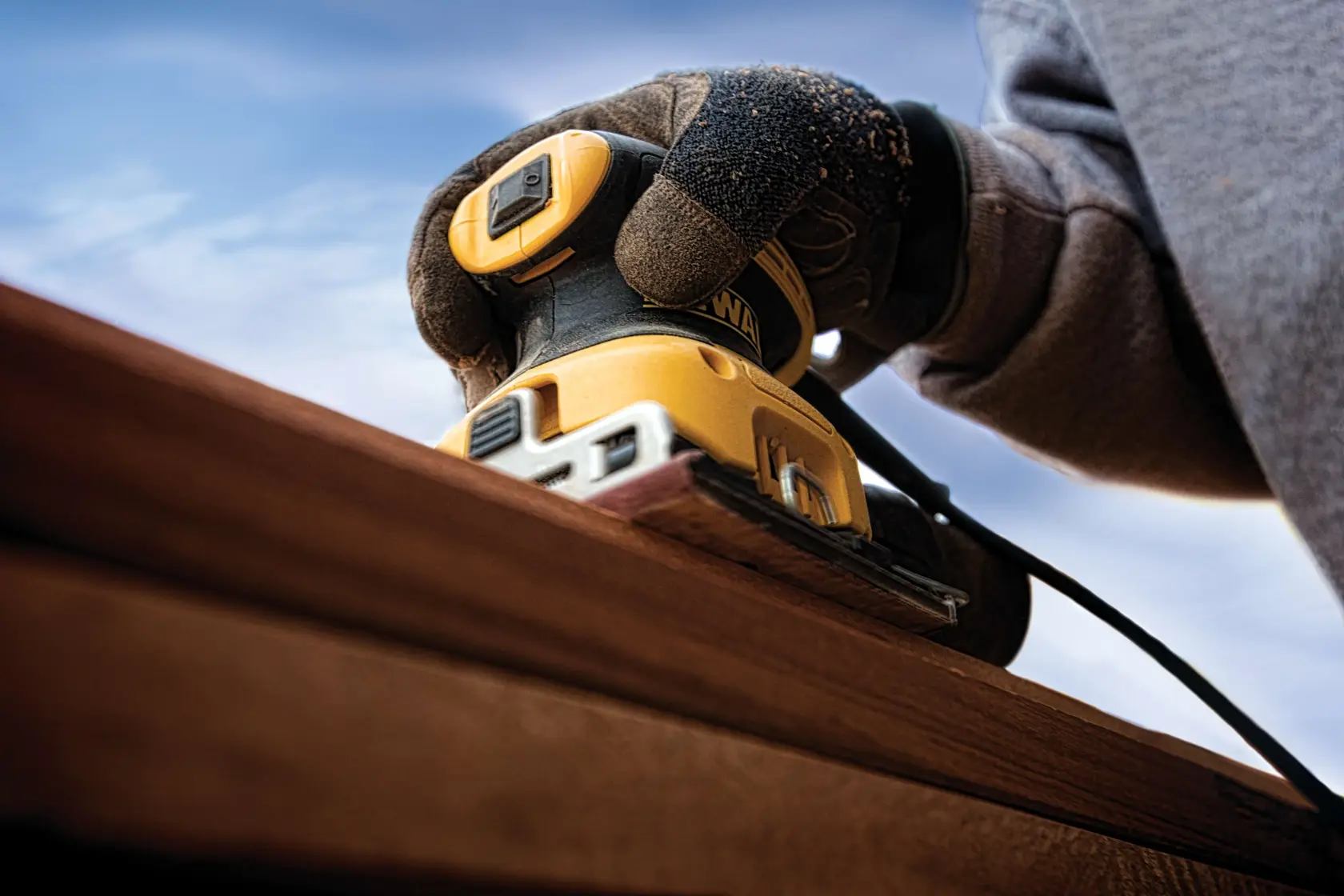 A close-up of a person using a DEWALT DWE6411 yellow and black electric sander to smooth a wooden surface outdoors. The person is wearing protective gloves.