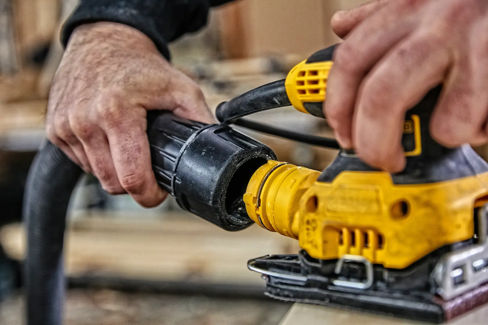 A person using a DEWALT electric sander on a wooden surface inside a workshop or garage. The sander is yellow and black, and the person's hand is holding it against the wood.